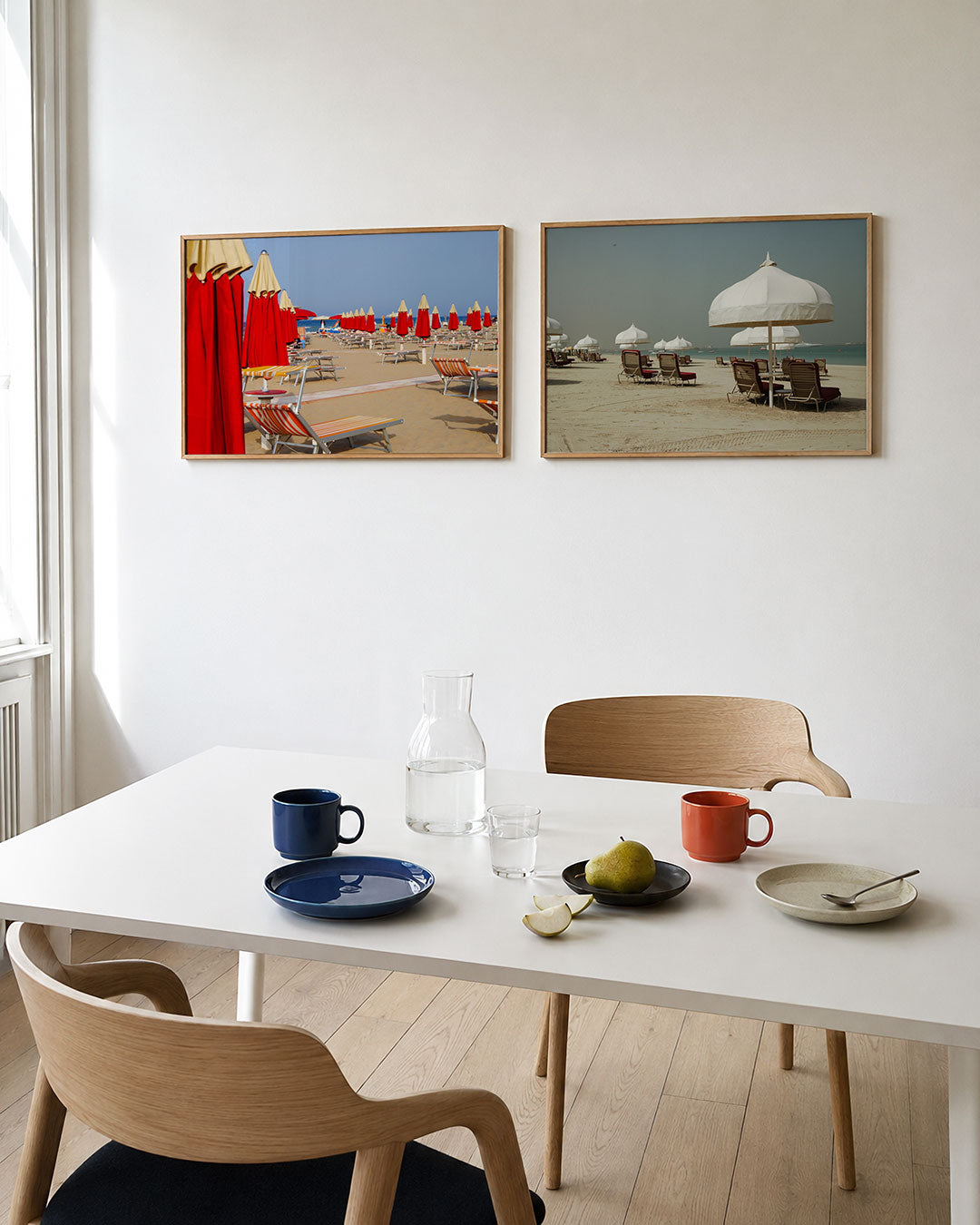 Twee liggende posters met rode parasols en ligbedden op een zonnig strand, aan de muur in een interieur